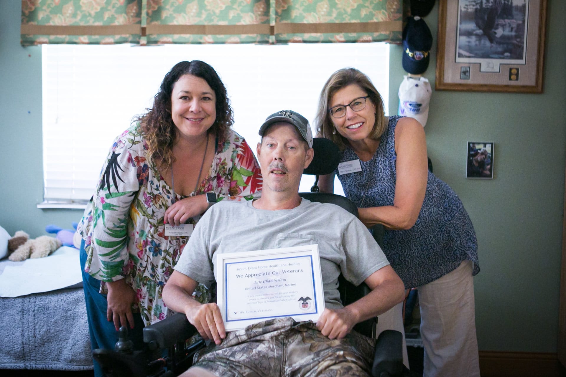 Two health care professionals with patient in wheel chair holding a "Veterans Appreciation" certificate.