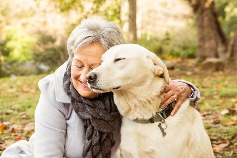 Senior woman smiling in the forest with her arms around her yellow Labrador dog